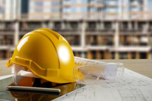 Image of a construction safety helmet and protective goggles sitting on an architectural drawing with a building site in the background